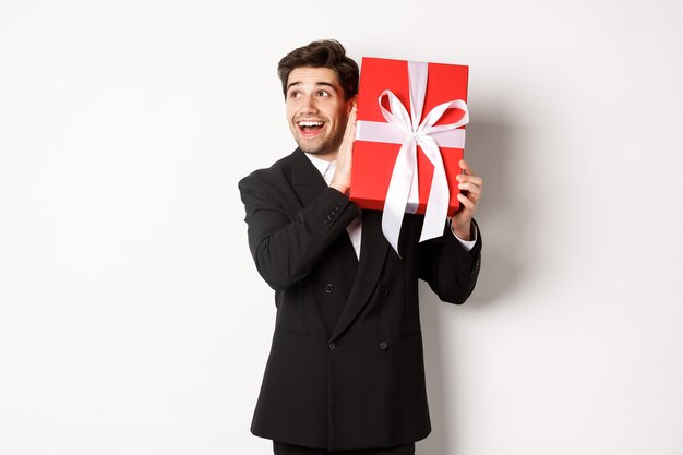 Imagen de chico guapo de ensueño en traje negro, caja de agitación con presente para preguntarse qué hay dentro, de pie contra el fondo blanco feliz.