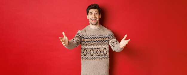Imagen de un chico guapo feliz y halagado con suéter navideño, alcanzando las manos para dar la bienvenida a los invitados en la fiesta de año nuevo, invitando a la celebración, de pie sobre un fondo rojo