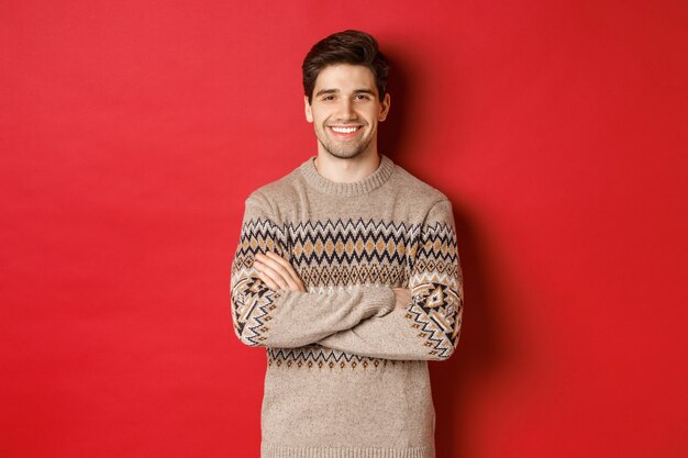 Imagen de chico guapo feliz en suéter de Navidad, sonriendo y mirando a cámara, celebrando las vacaciones de Navidad, de pie sobre fondo rojo.