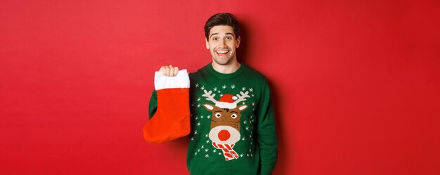 Imagen de chico guapo feliz en suéter sosteniendo calcetín de navidad y sonriendo divertido de pie contra...