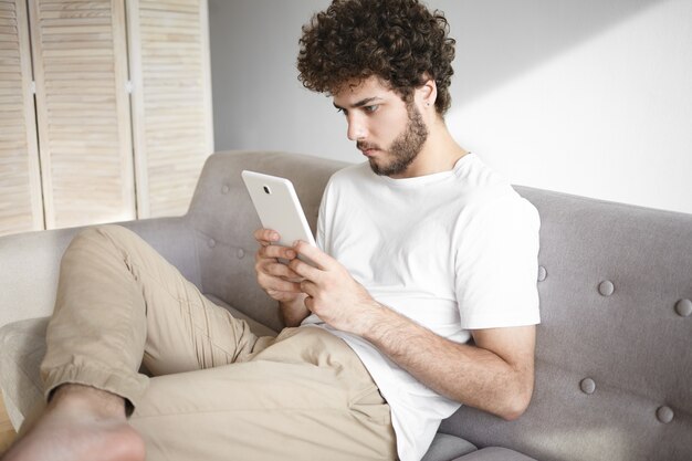 Imagen de un chico hipster concentrado con barba difusa y pies descalzos que pasan tiempo libre en el interior sosteniendo una tableta digital con ambas manos jugando videojuegos en línea, sentado cómodamente en un sofá gris