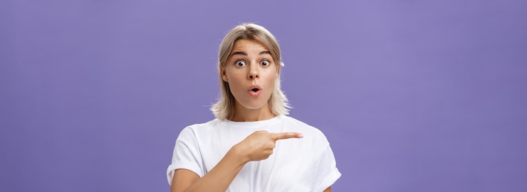 Foto gratuito imagen de la cintura de una mujer urbana abrumada, emocionada y sorprendida, atractiva, con camiseta blanca, los labios plegados, jadeando de asombro, siendo interrogada, haciendo preguntas y señalando a la izquierda sobre la pared púrpura.
