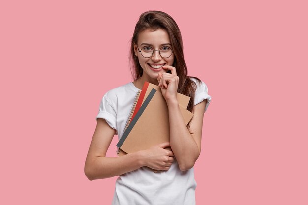 Imagen de una colegiala atractiva que mantiene el dedo cerca de la boca, sostiene el bloc de notas u organizador, vestida con una camiseta blanca informal, tiene el pelo largo peinado en un lado