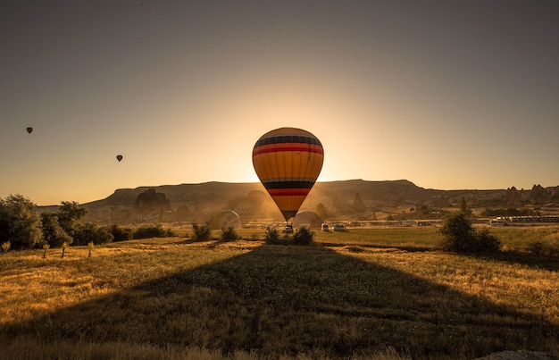 Imagen de un colorido globo aerostático en un campo rodeado de vegetación y montañas durante la puesta de sol