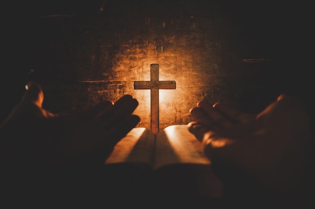 Imagen conceptual centrada en la luz de las velas con la mano del hombre sosteniendo una cruz de madera sobre la Biblia y el mundo borroso