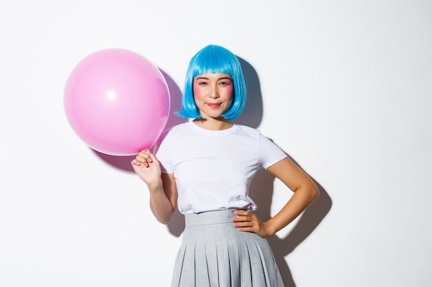 Imagen de coqueta mujer asiática descarada con peluca azul, vestida para la fiesta, sosteniendo un gran globo rosa y sonriendo confiada a la cámara.