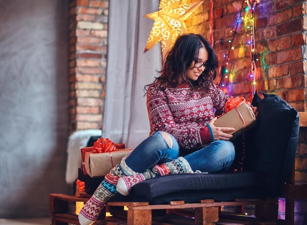 Imagen corporal completa de una mujer morena con anteojos vestida con jeans y un suéter rojo posando en un sofá de madera en una habitación con decoración navideña.