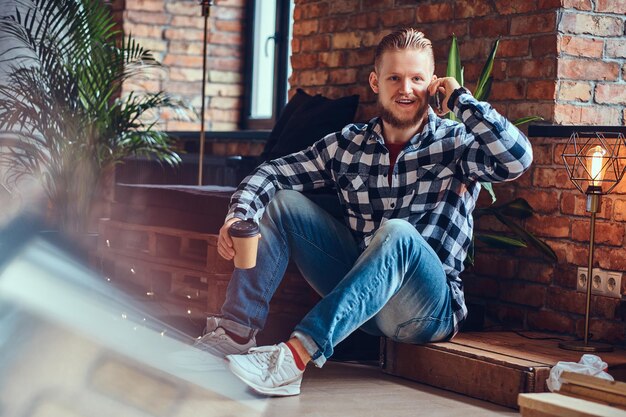 Imagen de cuerpo completo de un hombre elegante con barba rubia bebe café y usa un teléfono inteligente en una habitación con interior de loft.