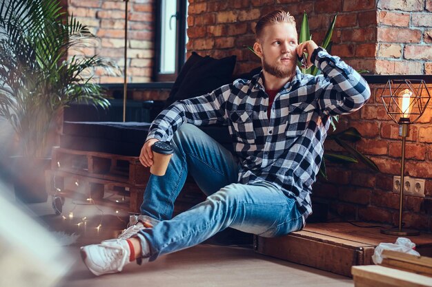 Imagen de cuerpo completo de un hombre elegante con barba rubia bebe café y usa un teléfono inteligente en una habitación con interior de loft.