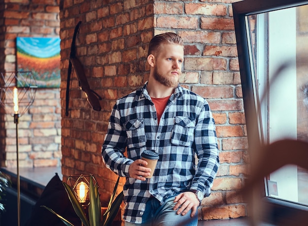 La imagen de cuerpo completo del hombre hipster rubio bebe café en una habitación con interior de loft.