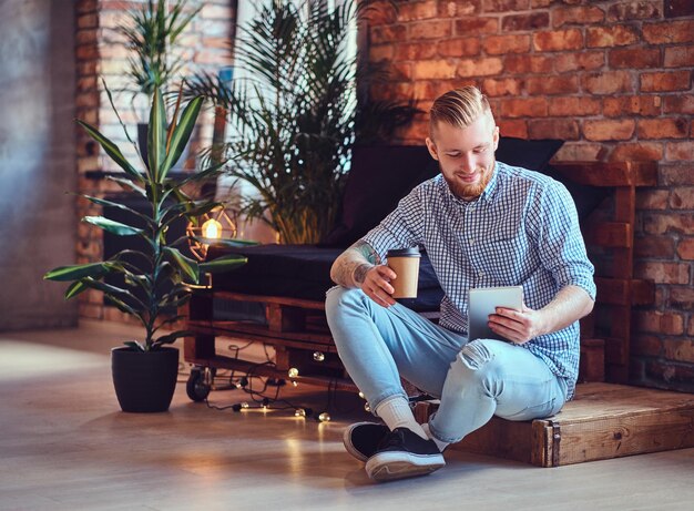La imagen de cuerpo completo de un hombre rubio y elegante vestido con una camisa de lana y jeans usando una tableta y bebe café en una sala de estar.