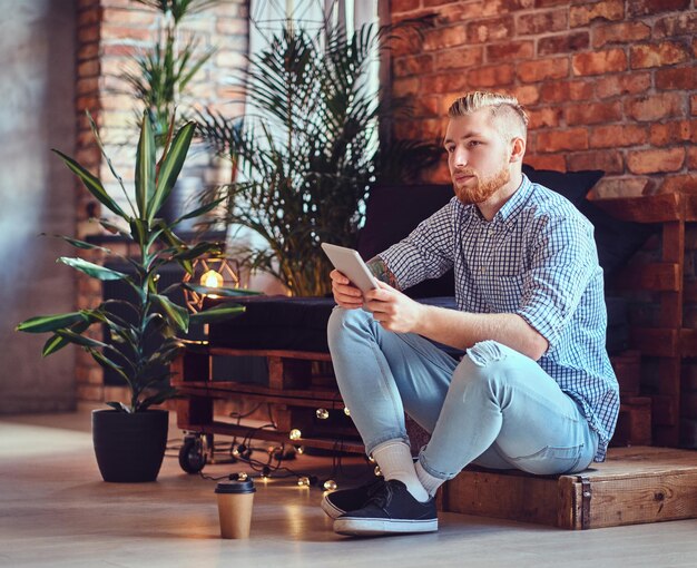 La imagen de cuerpo completo de un hombre rubio y elegante vestido con una camisa de lana y jeans usando una tableta en una sala de estar.