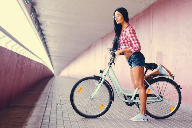 La imagen de cuerpo completo de la mujer deportiva con pantalones cortos de mezclilla y camisa polar posando en una bicicleta en un túnel con paredes rosas.