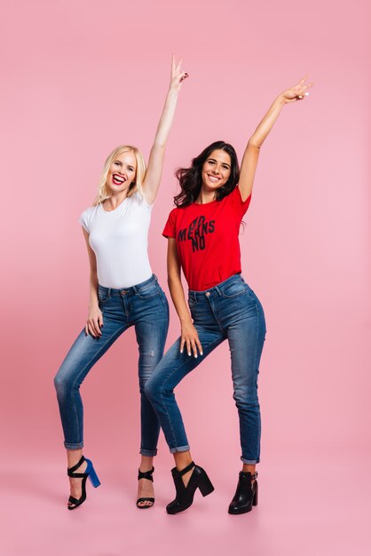 Foto gratuita imagen de cuerpo entero de dos mujeres sonrientes se regocijan en el estudio y mirando a la cámara en rosa