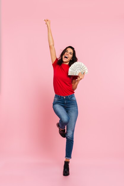 La imagen de cuerpo entero de la mujer morena feliz se regocija y sostiene dinero mientras mira a la cámara sobre rosa