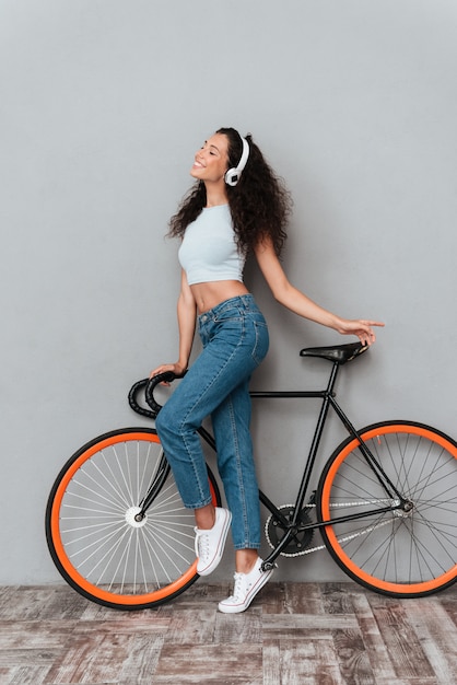 Imagen de cuerpo entero de mujer rizada sonriente de pie con bicicleta y escuchando música por los auriculares sobre fondo gris