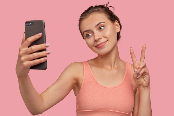 Foto gratuito imagen de una dama caucásica positiva con piel fresca, sostiene un teléfono inteligente moderno en el frente, hace un signo de paz, inclina la cabeza, vestida con modelos de chaleco informal sobre una pared rosa. chica guapa toma retrato selfie