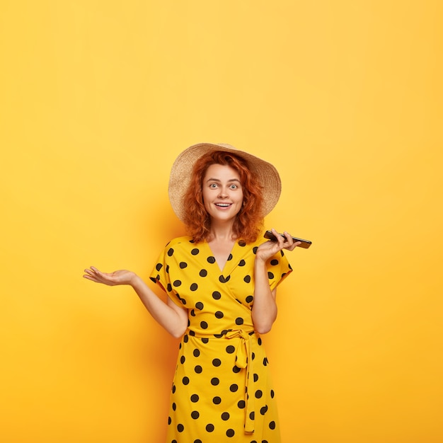 Imagen de la dama hermosa pelirroja sorprendida confundida levanta la palma, sostiene el teléfono móvil, se regocija de la nueva compra, usa sombrero de paja y vestido amarillo de lunares. Feminidad, estilo de vida