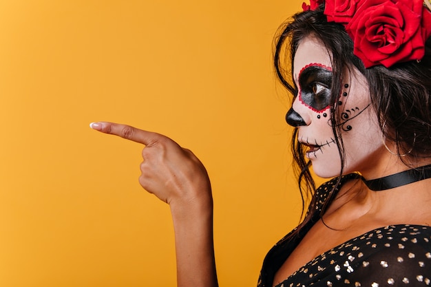 Imagen de dama en top negro de perfil. Chica con maquillaje de calavera en sorpresa muestra dedo a lado