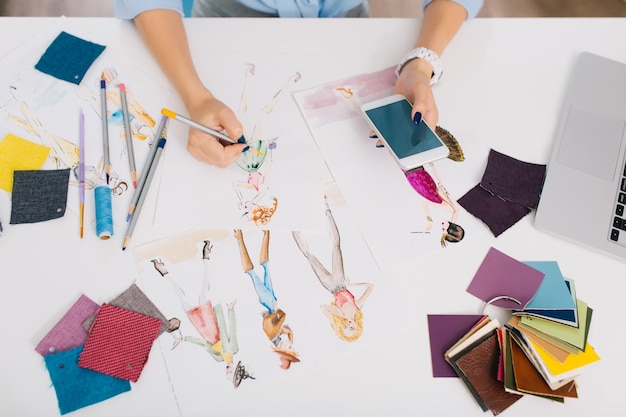Esta imagen describe los procesos de diseño de ropa. Hay manos de una niña dibujando bocetos con la ayuda de un teléfono en la mano. Hay un lío creativo con diferentes cosas sobre la mesa.