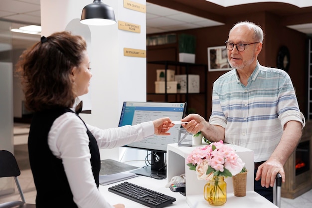 La imagen detallada muestra a un anciano caucásico jubilado recibiendo una tarjeta de acceso de una recepcionista en el mostrador de registro un turista masculino anciano dando la llave de la habitación a un empleado en la recepción del lobby del hotel