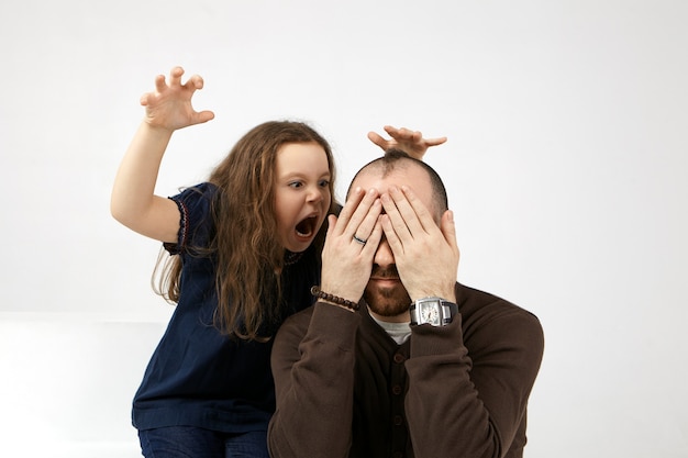 Foto gratuita imagen de una divertida niña europea vestida informalmente abriendo la boca ampliamente, gritando, asustando a su joven y elegante padre que está sentado y tapándose los ojos, sintiéndose asustado y aterrorizado