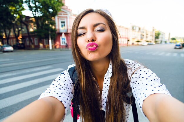 Imagen divertida de verano de una joven bonita viajera haciendo selfie en la calle, enviándote un beso, estado de ánimo positivo