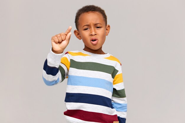 Imagen de un divertido colegial de piel oscura con un elegante jersey de rayas que tiene una expresión facial decepcionada, haciendo gestos con los dedos como si sostuviera algo muy pequeño. Descuento, venta y pequeño precio.