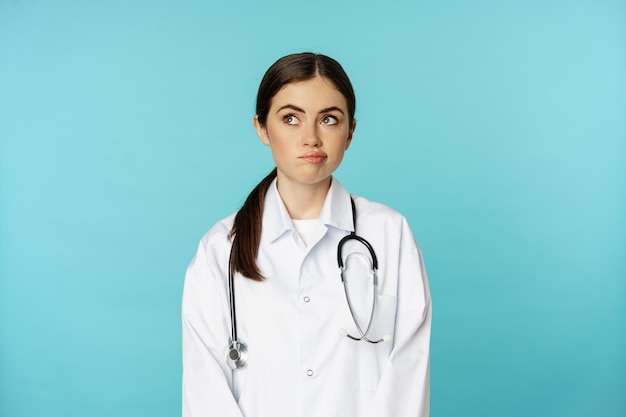 Foto gratuita imagen de una doctora, personal médico femenino con bata blanca de laboratorio, mirando hacia otro lado, tomando una decisión, pensando en algo, de pie sobre un fondo azul