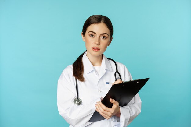 Imagen de una doctora profesional, médica con portapapeles escribiendo, escuchando a un paciente en una cita en la clínica del hospital, de pie sobre un fondo turquesa.