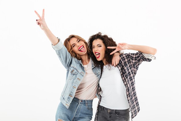 Imagen de dos chicas juguetonas de pie juntas y mostrando gestos de paz sobre una pared blanca