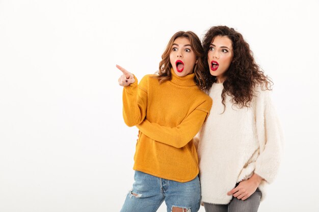 Imagen de dos muchachas sorprendidas en suéteres señalando y mirando a otro lado sobre bachground blanco