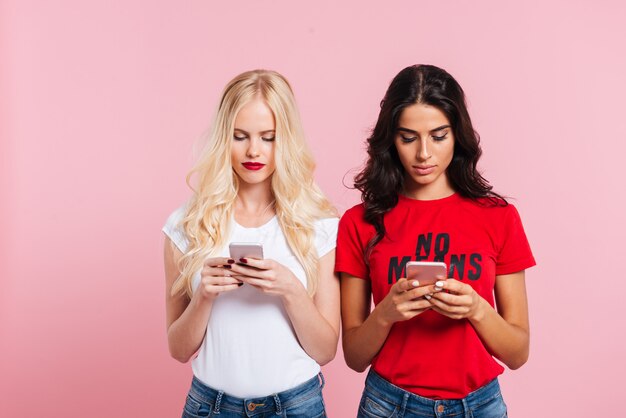 Imagen de dos mujeres bastante concentradas usando sus teléfonos inteligentes sobre rosa