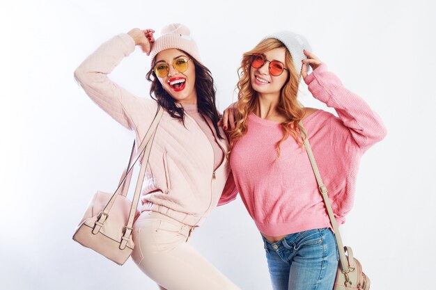 Imagen de dos niñas, amigas felices con ropa rosa elegante y un sombrero que deletrea divertido juntos. Fondo blanco. Sombrero y gafas de moda. Mostrando paz.