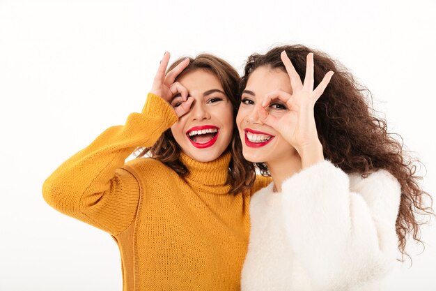 Imagen de dos niñas felices en suéteres divirtiéndose y mostrando gestos bien sobre la pared blanca