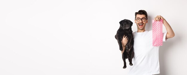 Imagen del dueño de una mascota hipster sosteniendo un lindo pug negro y una bolsa de caca de perro de pie sobre un fondo blanco