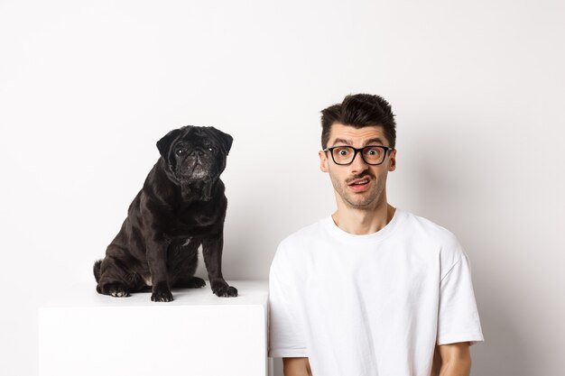 Imagen del dueño del perro inconformista sentado cerca de lindo pug negro, mirando a la cámara confundido y desconcertado, fondo blanco.