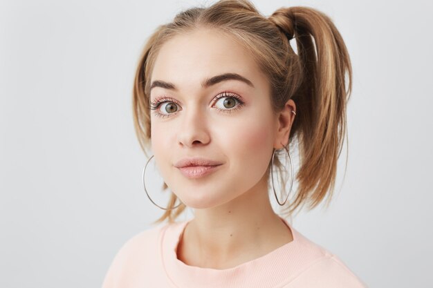 Imagen de una elegante adolescente de aspecto europeo con aspecto moderno, vestida con un suéter holgado de color rosa pastel, con dos coletas y aretes redondos, con un aspecto atractivo. Expresión facial