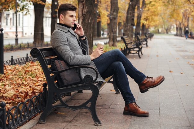 Imagen de un elegante hombre moreno con abrigo y jeans tomando café para llevar y hablando por teléfono inteligente, mientras está sentado en el banco en el parque