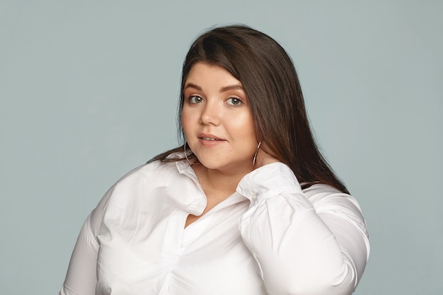 Foto gratuita imagen de elegante joven empleada con sobrepeso vestida con camisa blanca y grandes pendientes redondos tocando su cuello. mujer gordita hermosa aseada posando