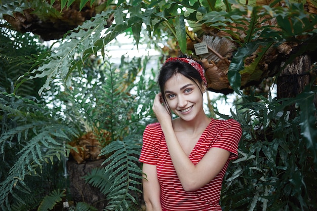 Imagen de elegante morena joven europea en vestido de moda y sombreros sintiéndose feliz y relajada de pie contra grandes hojas verdes de fondo de plantas exóticas, sonriendo ampliamente a la cámara