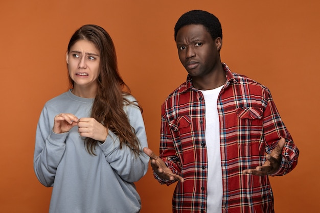 Imagen de emocional joven mujer de pelo largo con expresión de preocupación nerviosa mientras se siente culpable de pie junto a su confundido novio afroamericano que no entiende lo que está mal