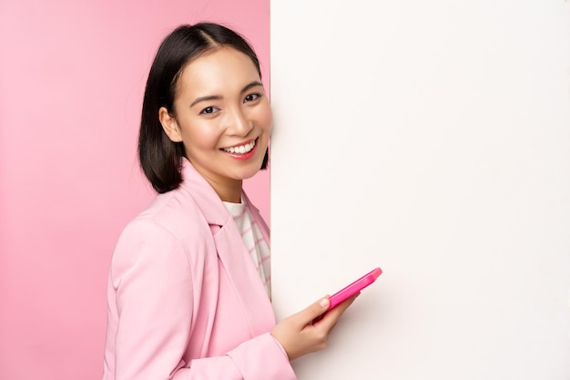 Imagen de una empresaria coreana en traje parada cerca de un anuncio de pared de información a bordo sosteniendo un teléfono inteligente y sonriendo posando sobre fondo rosa