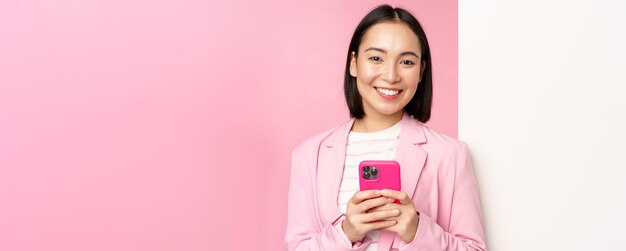 Imagen de una empresaria coreana en traje parada cerca de un anuncio de pared de información a bordo sosteniendo un teléfono inteligente y sonriendo posando sobre fondo rosa