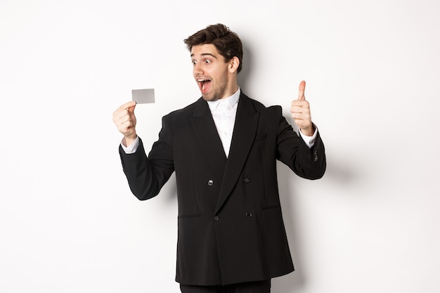 Imagen del empresario guapo emocionado, mostrando la tarjeta de crédito y el pulgar hacia arriba, de pie contra el fondo blanco.