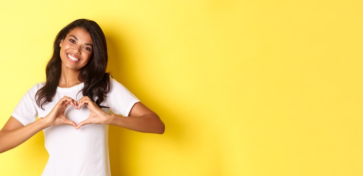 Foto gratuito imagen de una encantadora chica afroamericana con una camiseta blanca que muestra el signo del corazón y sonríe de pie junto a ti
