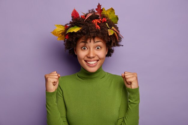 Imagen de la encantadora y feliz dama afroamericana aprieta los puños de alegría, sonríe ampliamente, disfruta del éxito, se siente contenta y llena de energía