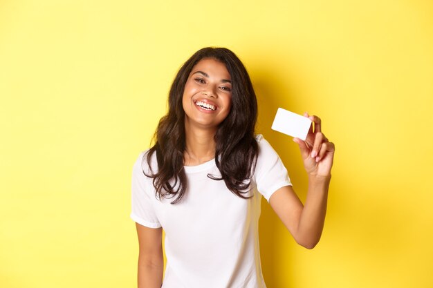 Imagen de la encantadora mujer afroamericana sonriendo feliz mostrando la tarjeta de crédito de pie sobre fondo amarillo