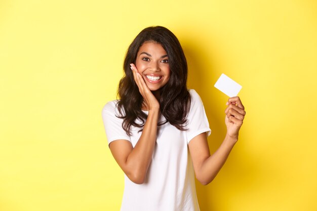 Imagen de la encantadora mujer afroamericana sonriendo feliz mostrando la tarjeta de crédito de pie sobre fondo amarillo