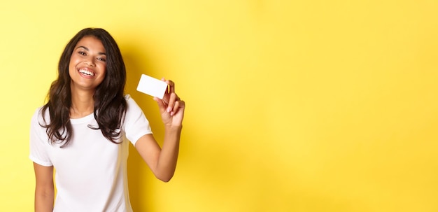 Foto gratuita imagen de una encantadora mujer afroamericana sonriendo feliz mostrando una tarjeta de crédito sobre un centrico amarillo